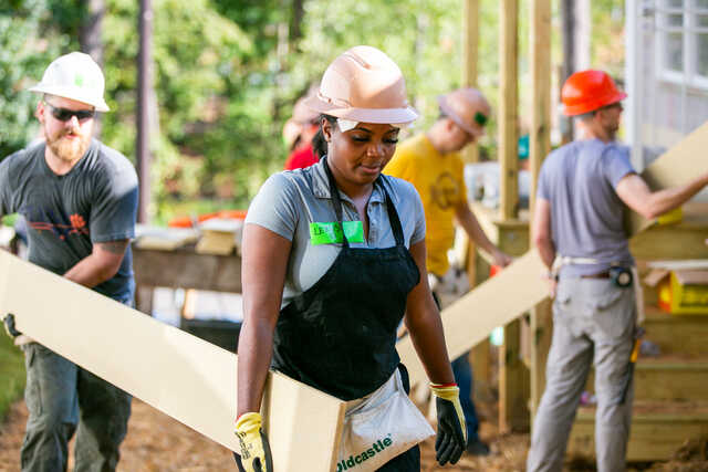 woman and other volunteers helping build a habitat house carrying wood wearing hard hats